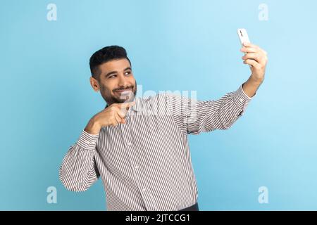 Ritratto di sorridente bel blogger uomo con barba puntando il dito alla fotocamera del telefono, facendo selfie o registrando video, indossando una camicia a righe. Studio in interni isolato su sfondo blu. Foto Stock