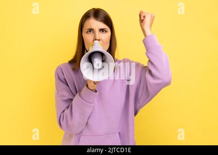Ritratto di donna che tiene il megafono vicino alla bocca, che parla forte, urla, che fa annuncio con il braccio sollevato, protestando, indossando la felpa con cappuccio viola. Studio al coperto isolato su sfondo giallo. Foto Stock