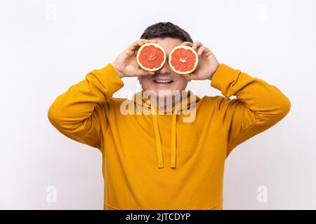 Uomo di mezza età che copre gli occhi con semi fettine di pompelmo e sorridente alla macchina fotografica, sano crudo concetto di cibo fresco vitamina, indossando felpa con cappuccio stile urbano. Studio in interni isolato su sfondo bianco. Foto Stock