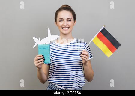 Ritratto di felice donna soddisfatta con acconciatura mordente indossando T-shirt a righe in piedi con bandiera tedesca e passaporto con aereo di carta, in viaggio. Studio in interni isolato su sfondo grigio. Foto Stock