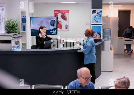 Receptionist usando il telefono fisso alla reception, dando documenti di rapporto al paziente da compilare prima di appuntamento di controllo medico. Lavoratore aiutare la donna con i moduli di consultazione, telefono cavo. Foto Stock