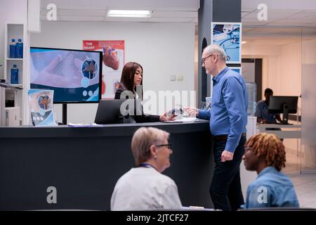 L'anziano paga la consultazione con la carta di credito, effettuando il pagamento elettronico alla reception dell'ospedale. Avendo appuntamento di controllo medico presso il centro sanitario, banco di registrazione. Foto Stock