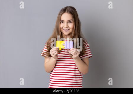 Bambina che indossa una T-shirt a righe che tiene e mostra due pezzi di puzzle, puzzle di corrispondenza e connessione, risolvere problemi con un buon risultato. Studio in interni isolato su sfondo grigio. Foto Stock