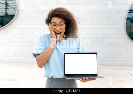 Eccitato scioccato stupito afro-americana giovane donna con capelli ricci, vestiti elegantemente, stand all'aperto, tiene un computer portatile aperto con bianco schermo mock-up in mano, guarda sorpreso la fotocamera Foto Stock