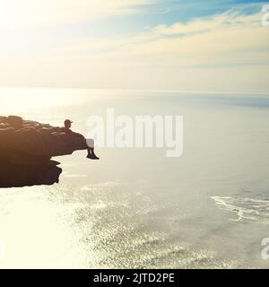 Seduto in cima al mondo, un giovane uomo non identificabile che ammira la vista dell'oceano da uno sbalzo di montagna. Foto Stock