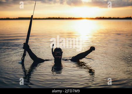 Subacqueo pesca di spearfishing festeggiando mentre sommerso in acqua di mare e tenendo un fucile da sparo. Allegro subacqueo godendo pesca spearfishing al tramonto. Foto Stock