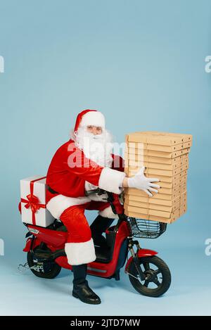 Consegna pizza uomo in costume di babbo natale in possesso di una grande pila di scatole di pizza mentre si siede al volante di uno scooter elettrico rosso a tutta lunghezza su Foto Stock