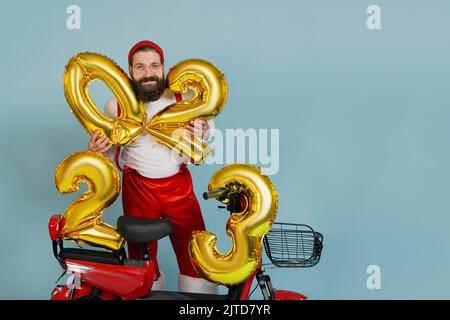 Un giovane uomo adulto in costume di babbo natale e sospensori tiene gonfiabili i numeri 2023 nelle sue mani sullo sfondo di uno scooter elettrico rosso su un iso blu Foto Stock