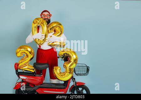 Giovane adulta hipster uomo in un costume di babbo natale e grandi occhiali a forma di cuore tiene gonfiabili numeri 2023 nelle sue mani su uno scooter elettrico rosso su un blu Foto Stock