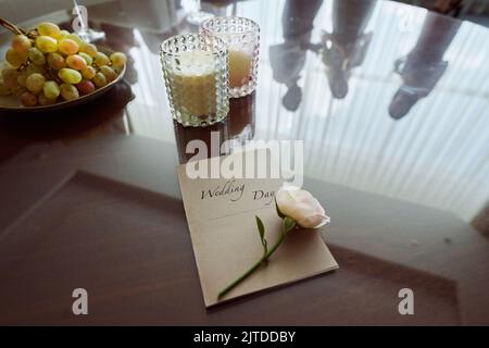 Invito di nozze con rosa bianca fresca, uva e due bicchieri con cocktail sul tavolo con il riflesso degli ospiti che brindano alla festa Foto Stock