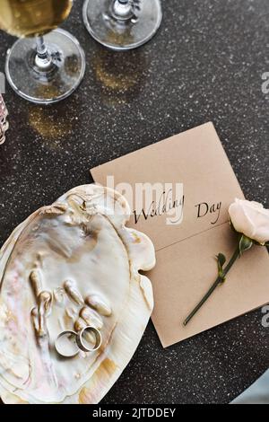 Flatlay di matrimonio composizione di conchiglia in perla con due anelli, invito, rosa bianca fresca e flauti con champagne sul tavolo Foto Stock