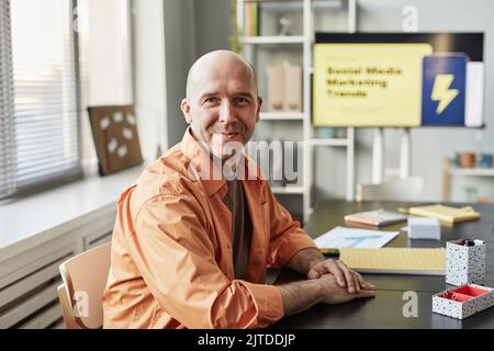 Ritratto di un uomo calvo sorridente che guarda la fotocamera mentre si siede sul posto di lavoro in un ufficio informale Foto Stock
