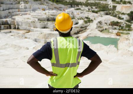 Vista posteriore del giovane caposquadra o ingegnere in giubbotto riflettente e casco di sicurezza in piedi sul territorio di vasta cava di marmo Foto Stock