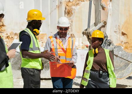 Gruppo di lavoratori interrazziali in hardhats e uniforme che discute il piano di lavoro alla riunione mentre il caposquadra fiducioso che indica il documento Foto Stock