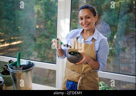 Donna ispirata fiorista sorride guardando la macchina fotografica, tiene un rosmarino in vaso e pala da giardino con terreno fertilizzato in mano Foto Stock