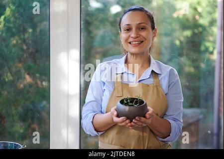 Bella donna, fiorista ispirato in grembiule beige tenendo in mano un rosmarino in vaso e sorridendo, guardando la macchina fotografica Foto Stock
