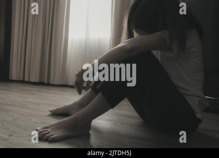Donna depressa e stressata seduta sul pavimento vicino alle tende in camera da letto. Donna triste seduta nella stanza buia. Ragazza con problemi di salute mentale. Foto Stock