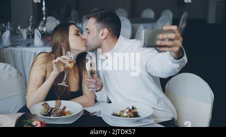 Coppia amatevole sta prendendo selfie con i calici dello champagne usando lo smartphone mentre sta avendo cena romantica nel ristorante. Stanno sorridendo, baciando e posando guardando la macchina fotografica. Foto Stock