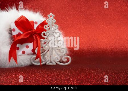 Scatola regalo bianca con puntini rossi nel cappello di Babbo Natale e albero di Natale su sfondo rosso brillante. Anno nuovo. Spazio di copia Foto Stock