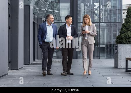Gruppo di lavoro diversificato, tre lavoratori maschi e femmine che camminano e chiacchierano discutendo piani, al di fuori dell'edificio degli uffici Foto Stock