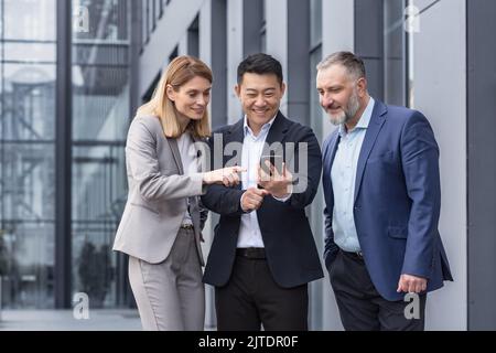 Gruppo di lavoro diversificato, tre lavoratori di sesso maschile e femminile al di fuori dell'edificio ufficio allegro sorridente e felice guardando video al telefono Foto Stock