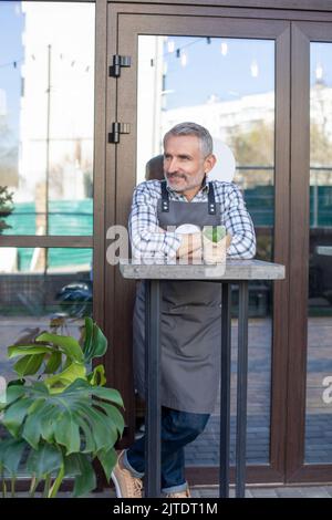 Sorridente dipendente di Street cafè che si trova all'aperto e guarda lontano Foto Stock