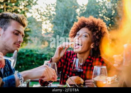 Bella ragazza afroamericana che vive uno stile di vita sano e sorridente e scherzando mentre mangia e beve vino rosso con i suoi amici al pub all'aperto r Foto Stock
