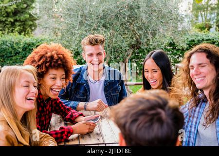 Gruppo di giovani seduti in un bar e sorridente e guardando theyr smartphone - felice gruppo multietnico di amici che parlano utilizzando smartphone in pub Foto Stock