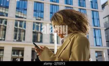 Donna afro-americana cammina a scrivere messaggi al ragazzo su smartphone. La giovane signora dai capelli cinky legge le notizie sorridendo mentre cammina davanti all'edificio dell'ufficio Foto Stock