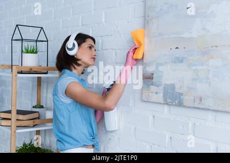 brunette donna in cuffie wireless pulizia foto con straccio e detergente Foto Stock