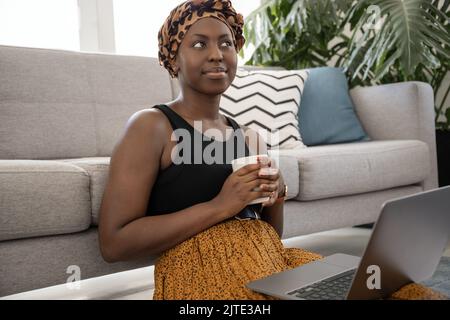 Bella giovane gen z nero africano donna, indossando tradizionale sciarpa testa, seduto sul piano salotto tenendo una tazza di caffè e computer portatile in giro Foto Stock