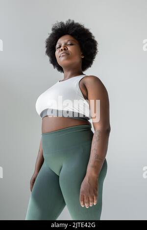 vista dall'angolo basso di una donna afro-americana in tatuata più misura in abbigliamento sportivo isolato su grigio, immagine stock Foto Stock