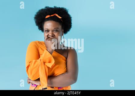preoccupato afro americano corpo donna positiva in abito arancione isolato su blu, immagine stock Foto Stock