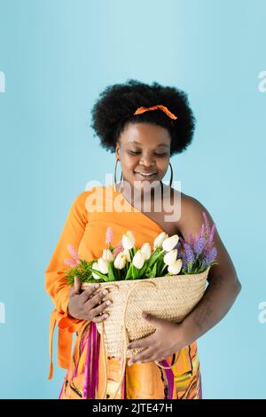 sorridente corpo afro-americano positivo donna che tiene sacchetto di paglia con fiori isolati su blu, immagine stock Foto Stock