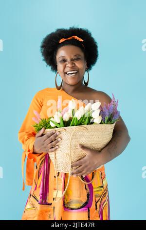 gioioso corpo africano americano positivo donna che tiene sacchetto di paglia con fiori isolati su blu, immagine stock Foto Stock