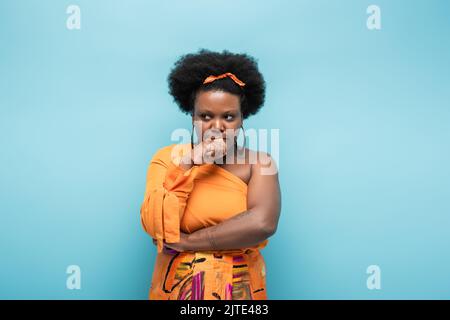stressato corpo afro-americano positivo donna in abito arancione e orecchini cerchio isolato su blu, immagine stock Foto Stock