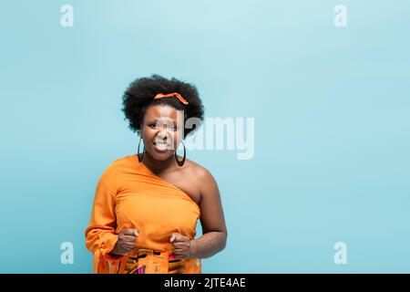 preoccupata african american corpo positivo donna in orecchini cerchio e abito arancione isolato su blu, immagine stock Foto Stock
