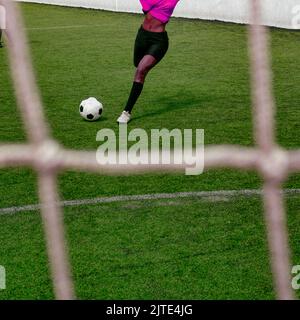 Irriconoscibile donna di colore scuro con pelle in una divisa rosa viola da calcio sport calcio a calci una palla di calcio in gol sul campo di calcio. Foto Stock