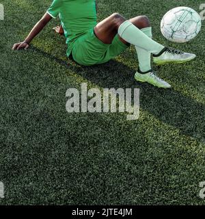 Uomo dai colori scuri e irriconoscibile in un'uniforme da calcio verde che regge una palla da calcio mentre si sdraia sul campo da calcio. Foto Stock