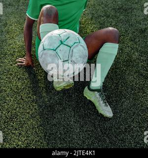 Uomo dai colori scuri e irriconoscibile in un'uniforme da calcio verde che regge una palla da calcio mentre si sdraia sul campo da calcio Foto Stock