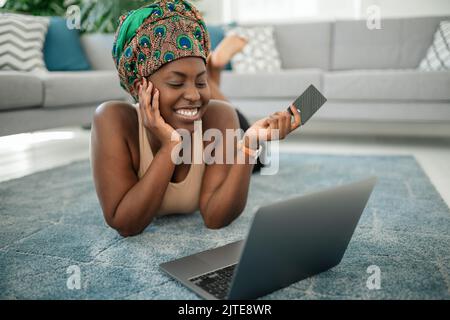 Donna africana nera che indossa il tradizionale velo, facendo acquisti online utilizzando il laptop e la carta di credito. Stendendo sul tappeto nel salotto a casa Foto Stock