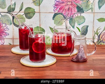 Tè al ghiaccio al lampone in una speciale teiera di vetro, con due bicchieri da tè al ghiaccio e cannucce in borosilicato, su un tavolo in noce e piastrelle in ceramica sullo sfondo. Foto Stock
