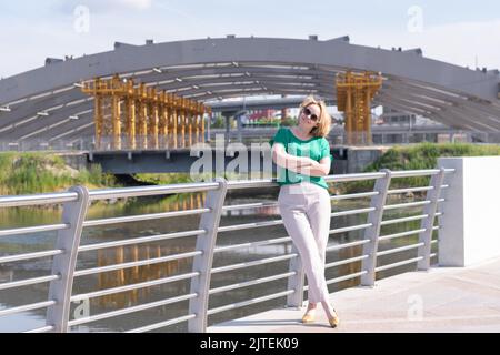 Un ritratto di una ragazza in una blusa verde, pantaloni e occhiali da sole sullo sfondo di architettura urbana incompiuta. Una ragazza cammina intorno ad una nuova zona di Foto Stock