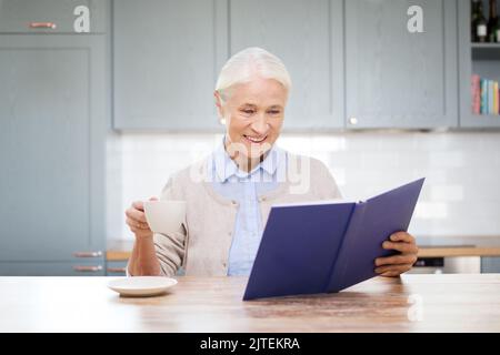 felice donna sorridente che legge il libro in cucina Foto Stock