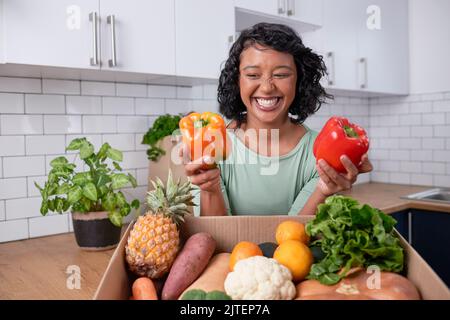 Una giovane donna multietnica tiene due peperoni dolci fuori dalla scatola di consegna di verdure fresche Foto Stock