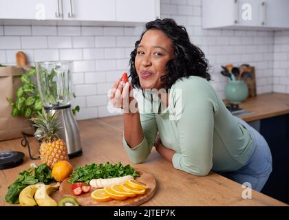 Una giovane donna attraente assaggia una fragola mentre prepara la frutta per il frullato Foto Stock