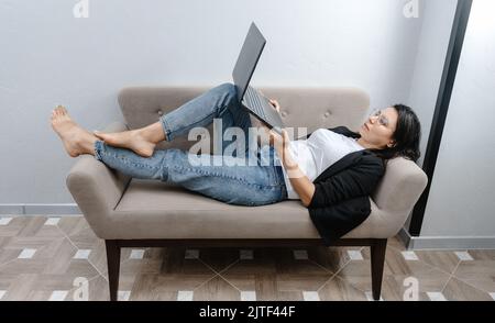 Una donna felice e tranquilla che lavora al suo laptop nella comodità del suo divano. Lavoro a casa. Lavorare comodamente al computer. Foto Stock