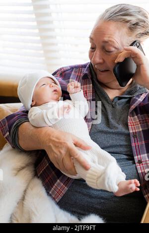 Nonna che tiene in mano il nipote neonato (0-1 mesi) e che parla al telefono Foto Stock