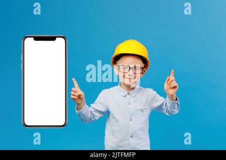 Carino ragazzo di costruzione in elmetto giallo in piedi in studio interno su sfondo blu e la direzione di puntamento con le dita vicino enorme phon mobile Foto Stock