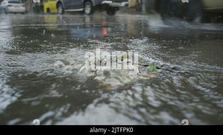 Un flusso di acqua piovana scorre nel tempesta drenaggio auto sono in guida in background. Forte pioggia torrenziale in città. Foto Stock